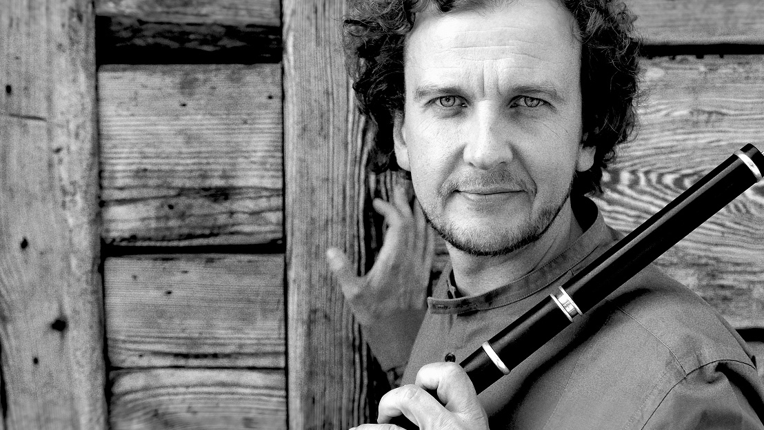 A man holds a brass instrument in one hand, the other hand rests on a wooden wall. The man smiles, looking directly into the lens.