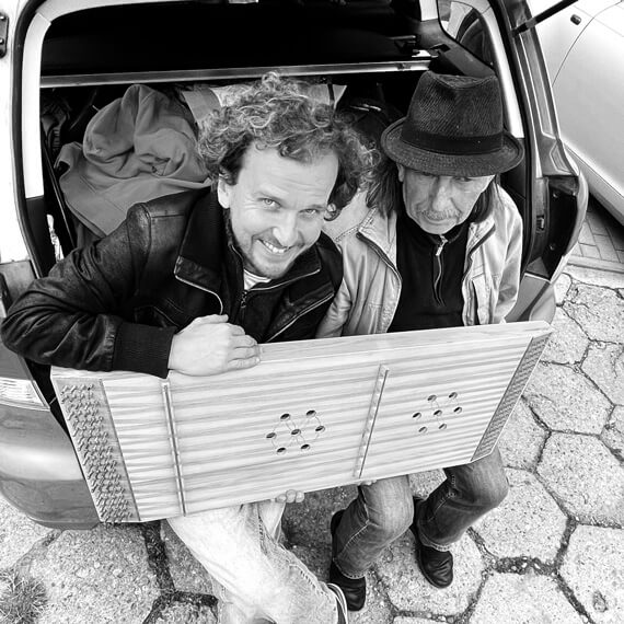 Dwóch mężczyzn siadzi w otwartym bagażniku samochodu. Jeden z nich trzyma na kolanach instrument